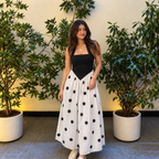 A woman stands between two potted leafy plants wearing a sleeveless black top and a long white skirt with large black polka dots. The background is a light beige wall and gray floor.