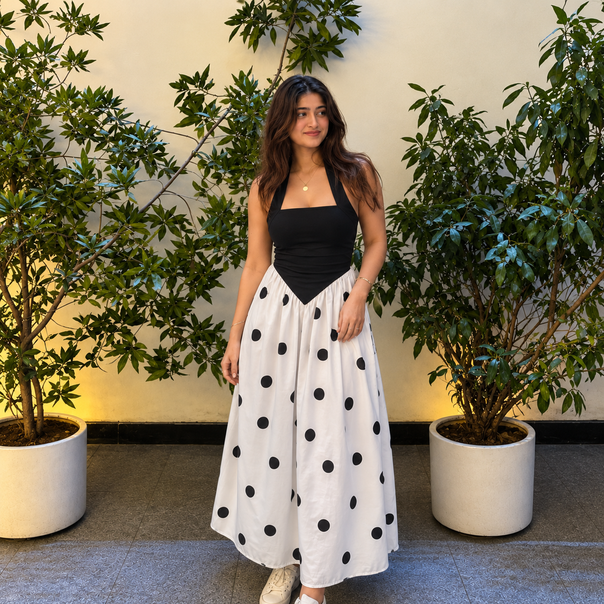 A woman stands between two potted leafy plants wearing a sleeveless black top and a long white skirt with large black polka dots. The background is a light beige wall and gray floor.